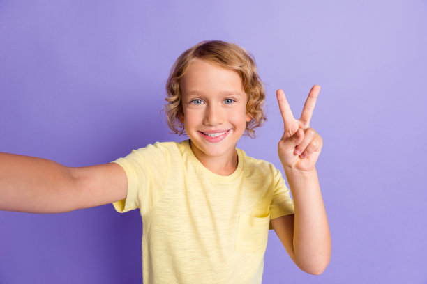 正面男孩的肖像自拍照片，使v标志穿黄色t恤孤立在紫罗兰色背景图片下载