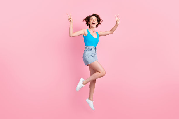 漂亮迷人的运动的年轻女士的全身尺寸照片微笑，张开嘴，跳，举起手，展示v字，穿牛仔迷你裙，蓝色背心，运动鞋孤立的粉色背景图片下载