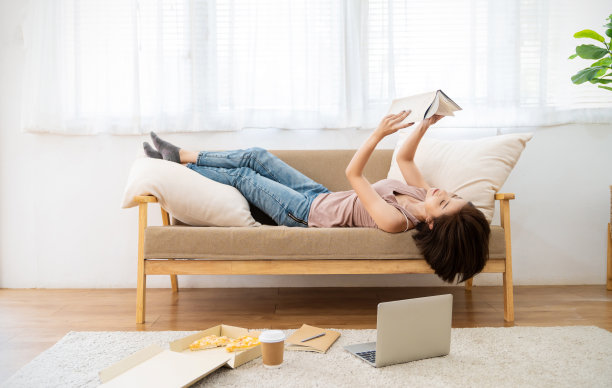 年轻的亚洲女人阅读放松在家里的沙发上。年轻的亚洲女孩订披萨外卖，在家工作。图片下载