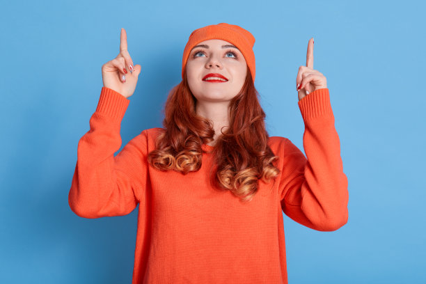 有魅力的女人与红色波浪发指向上方的拷贝空间区域，显示你的广告，穿着休闲衣服和帽子，孤立在蓝色背景。图片下载