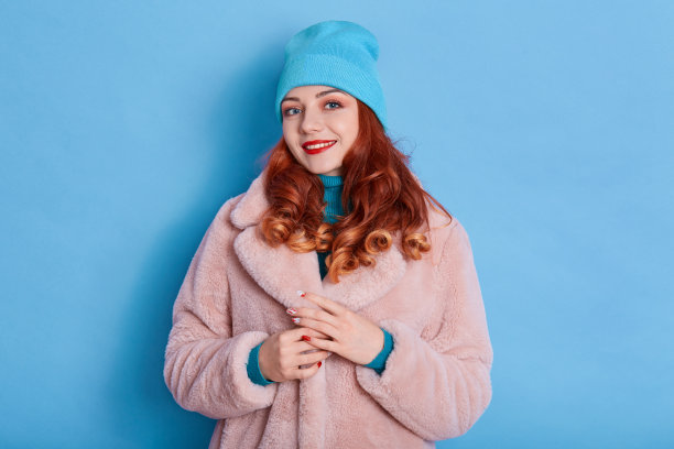 漂亮的女人穿着粉红色的皮草大衣，戴着蓝色的帽子，露出牙齿般的微笑，涂着红色的口红，看着镜头，有红色卷发的女性，看着镜头，表达积极的情绪。图片下载
