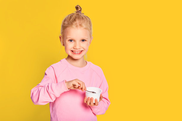 可爱的小女孩吃美味的酸奶在黄色的背景。4-5岁的孩子穿着粉红色运动衫在工作室摆姿势。空间的文本图片下载