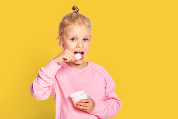 可爱的小女孩吃美味的酸奶在黄色的背景。4-5岁的孩子穿着粉红色运动衫在工作室摆姿势。空间的文本图片下载