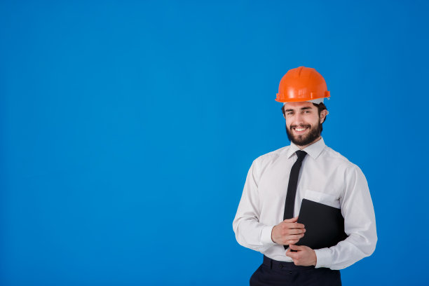 一名留着胡子的年轻男性建筑工人，身穿白色衬衫，戴着建筑头盔，背景是蓝色。工业工程师-建筑师在一个孤立的背景上拿着带有文件的剪贴板图片下载