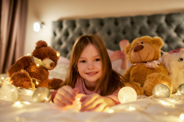 一个可爱的小女孩独自躺在一张大床上，光着脚看着镜头，微笑着，身边有她的泰迪熊和花环图片下载