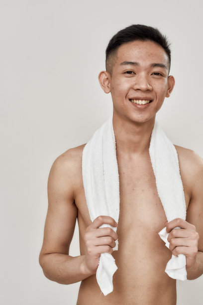 赤裸上身的年轻亚洲男子的肖像，有问题的皮肤和色素沉着在他的脸摆姿势与毛巾周围他的脖子孤立在白色的背景。美容、护肤的常规概念图片下载