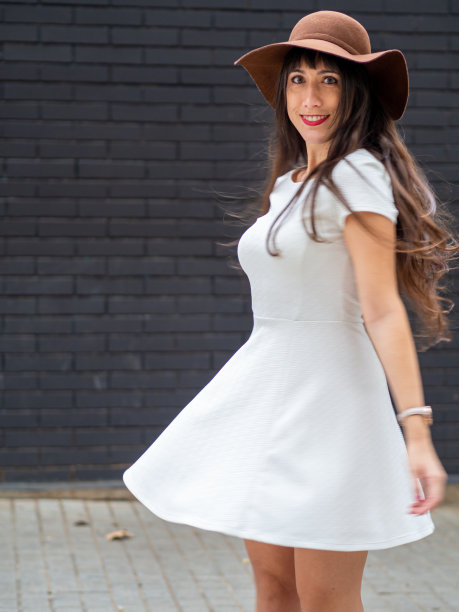年轻的女子穿着白色的裙子和帽子在一堵黑色的砖墙前跳舞。图片下载