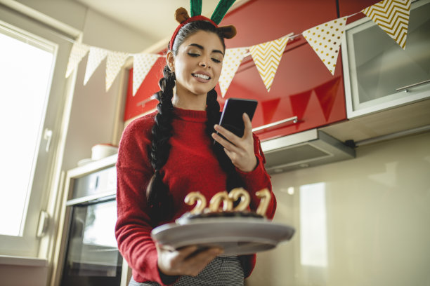 美丽的女人庆祝圣诞节，有视频通话，拿着巧克力蛋糕的蜡烛形状的数字2020，代表即将到来的新年图片下载