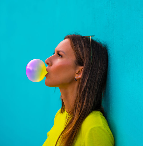 年轻美丽的女士在黄色t恤站在孤立的蓝绿色背景墙，而吹泡泡彩色口香糖肖像。图片下载