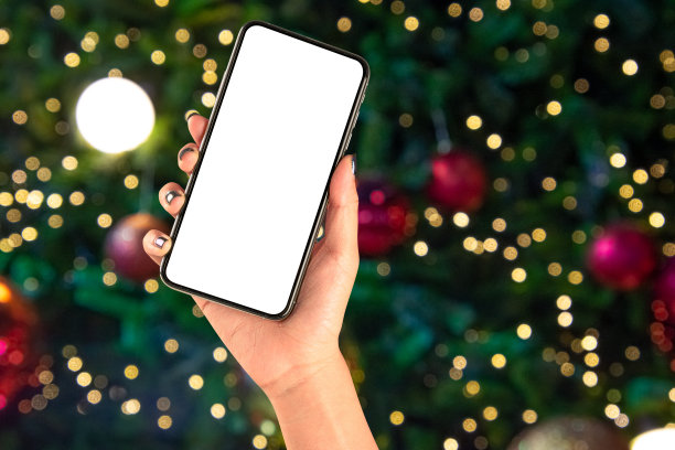 近距离手持手机孤立在白色背景与照明模糊的散景圣诞树装饰与悬挂灯球。图片下载