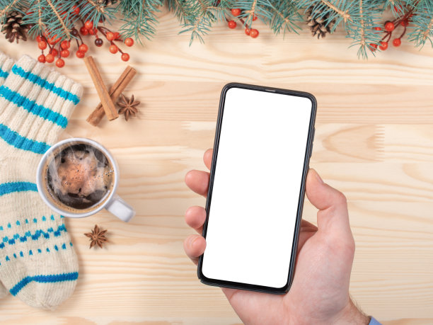 圣诞背景上，一个男人的手拿着黑色手机的黑屏模型。手持智能手机，在模糊的圣诞装饰背景上模拟圣诞季。图片下载