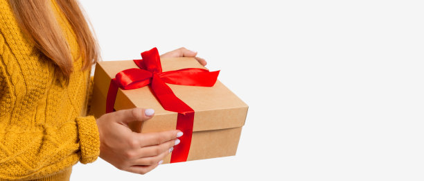 女性手中正拿着用红色蝴蝶结装饰的工艺品礼盒圣诞、新年。孤立的副本空间。一个穿着舒适温暖的黄色毛衣没有脸的女人。光背景横幅图片下载