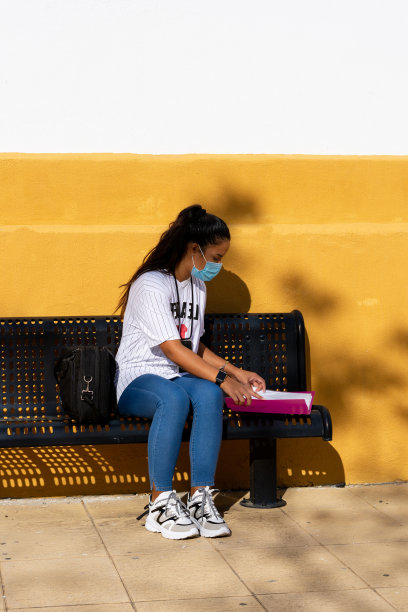 一个大学女生坐在长凳上学习，墙上涂着黄色，文件夹是粉红色的图片下载