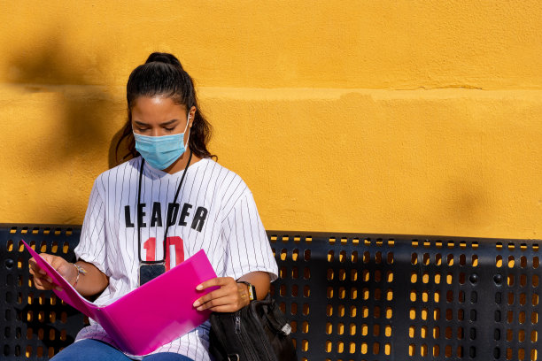 一个大学女生坐在长凳上学习，墙上涂着黄色，文件夹是粉红色的图片下载