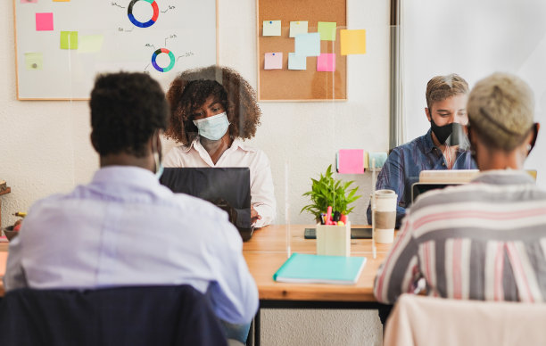 年轻的多种族人群戴着防护口罩，用塑料分隔物在办公室工作——关注非洲女孩图片下载