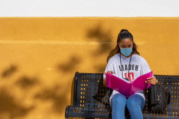 一个大学女生坐在长凳上学习，墙上涂着黄色，文件夹是粉红色的图片下载