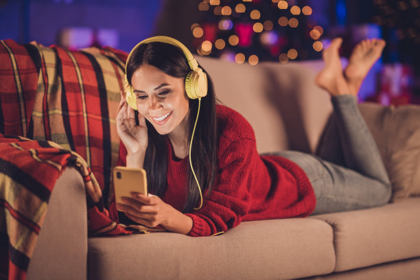 照片中的女孩躺在沙发上触摸耳机，一只手拿着电话，穿着红色的套头衫在室内装饰圣诞节图片下载