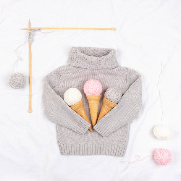 灰色针织毛衣上放着三个毛线冰淇淋和蛋筒，白色背景上放着木制的针织针。编织，爱好和手工概念。平铺图片下载