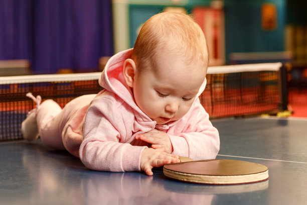 一个6个月大的白人婴儿穿着粉红色的衣服，正趴在网球桌上。一个小女孩拿着一个乒乓球拍图片下载