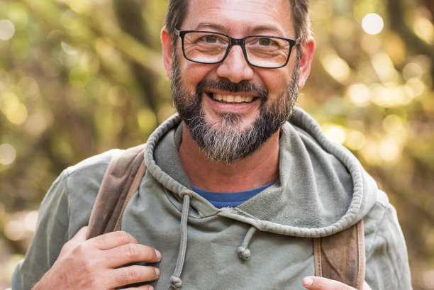 肖像中的成年男子微笑着看着相机与散焦绿色的自然背景-人与户外休闲活动-英俊的男子戴着眼镜和胡子图片下载