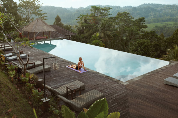 早上在印度尼西亚巴厘岛的泳池边做瑜伽的美丽女孩。年轻苗条的女人在运动服站在眼镜蛇姿势或布jangasana运动垫附近的无限泳池对热带景观。图片下载