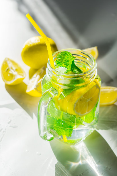 将柠檬和薄荷水倒入一个透明的杯子里，放在白色的桌子上，上面放着柠檬片，在早晨明亮的阳光下闪闪发光。垂直图图片下载