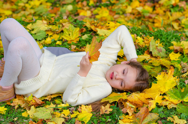 一个穿着白色针织连衣裙的小女孩微笑着躺在落叶上。秋天公园里明亮的黄色、橙色的枫叶。图片下载
