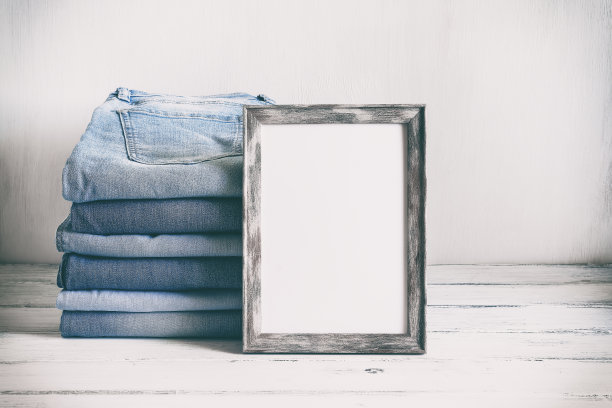 空白的相框模型和蓝色牛仔裤堆在一张旧的白色木桌上。模板为您的现代设计。复古风格的照片色调。有选择性的重点。图片下载