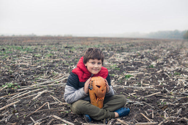 十几岁的男孩在温暖的衣服坐着，并持有万圣节雕刻的南瓜杰克-o'-灯在田野素材图片