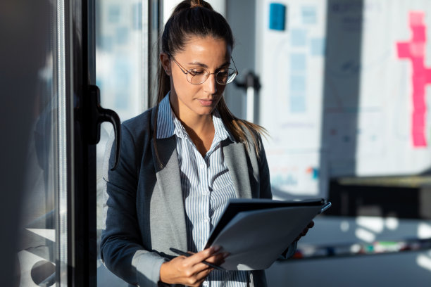 这张照片拍摄的是一位年轻女子站在办公室的窗户旁边，专心致志地阅读一些文件。图片下载