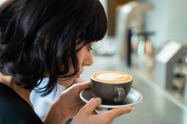 美丽的女性白人顾客在室内咖啡馆餐厅拿着一杯拿铁艺术咖啡。那个女人看着咖啡，闻着咖啡的芳香，幸福地微笑着。饮料喝的概念图片下载