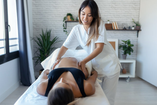 美丽的年轻理疗妇女按摩肚子上的孕妇在担架上在家。图片下载