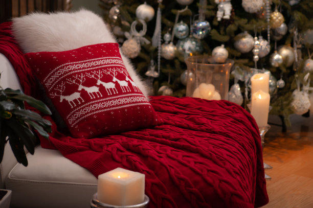 华丽的红色针织毯子和沙发上的鹿图案垫子，地板上的大蜡烛和装饰的圣诞树在背后的圣诞静物图片下载