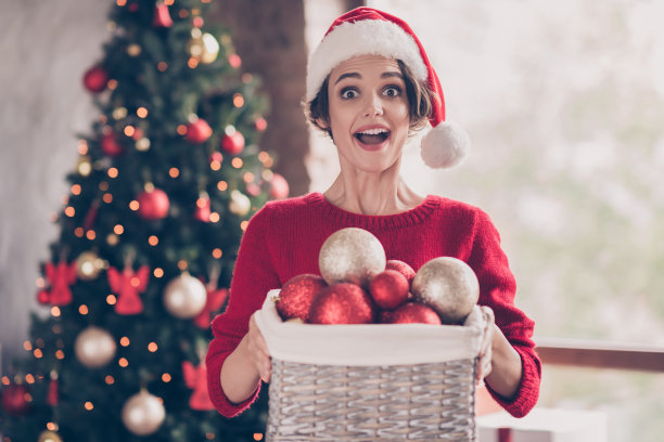 可爱的照片兴奋的女士拿着新年盒子球玩具张着嘴戴着圣诞老人的头饰红色套头衫装饰室内客厅图片下载