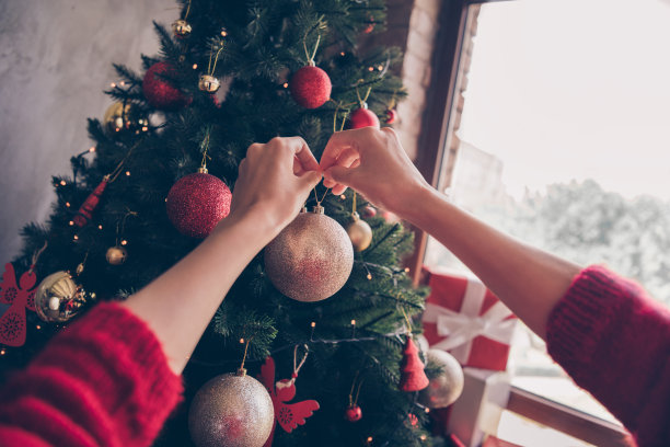 小女孩的裁剪照片，手挂装饰品，铃铛，玩具，金球，常青树，穿红色套衫装饰客厅室内图片下载