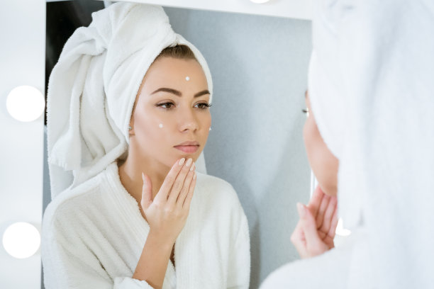 一个快乐的年轻女子在镜子前用毛巾敷面霜，这是在家护肤的概念图片下载