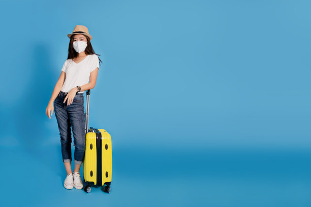 年轻的亚洲女性游客戴着面具和夏季休闲装与黄色手提箱。她在蓝色背景下很快乐。图片下载