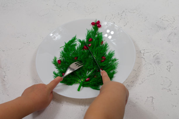 这棵树衬着绿色的莳萝叶子，用白色盘子上的红石榴果装饰。孩子们的手拿着刀叉。新年的食物。图片下载
