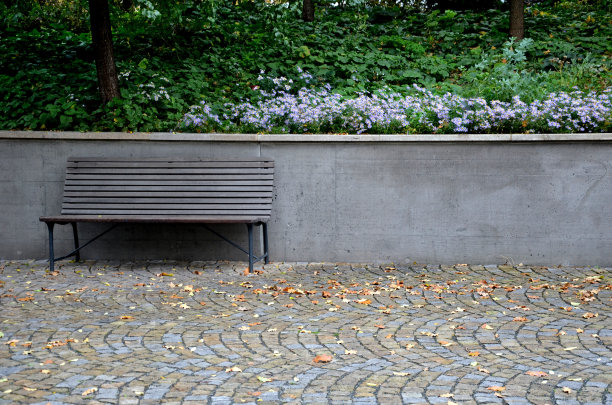 公园内的灰色支撑混凝土墙旁的长椅。紫苑在花坛的墙上开着花。木镶板公园长椅与金属黑色框架。图片下载