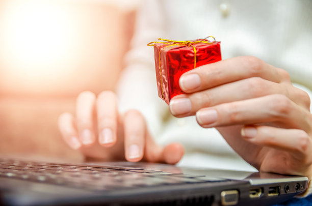 在特写镜头中，一个穿着白色毛衣的女人把笔记本电脑放在膝盖上，手里拿着一个红色的礼品盒。照片与照明。圣诞节、新年、节日的概念图片下载