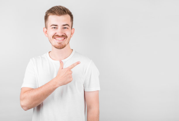 年轻英俊的男子穿着休闲衬衫站在白色的背景，微笑的脸指向与手和手指向上的一边，脸上的快乐和自然的表情图片下载