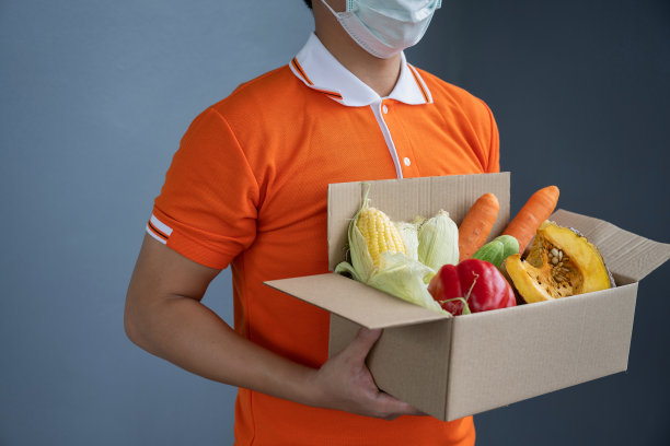一名身穿橙色制服的亚洲送货员戴着口罩，搬运装着食品、杂货、水果的箱子，站在房前。新冠肺炎期间的邮差及快递杂货送货服务。图片下载