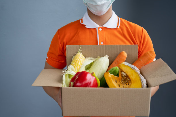 一名身穿橙色制服的亚洲送货员戴着口罩，搬运装着食品、杂货、水果的箱子，站在房前。新冠肺炎期间的邮差及快递杂货送货服务。图片下载