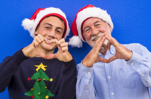 两位快乐的圣诞老人，一位爷爷和他十几岁的孙子，微笑着用双手做了一个心形。家庭、爱和乐趣的概念图片下载