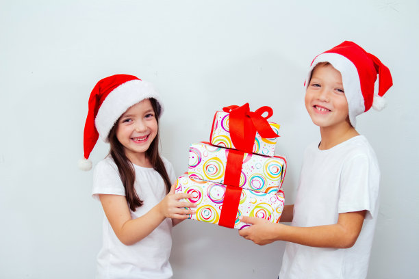 圣诞节的孩子。一个戴着红色圣诞老人帽、穿着白色t恤、面带微笑的男孩和女孩组成了一个大礼盒，孤立在灰色的背景下。圣诞快乐，礼物大减价图片下载