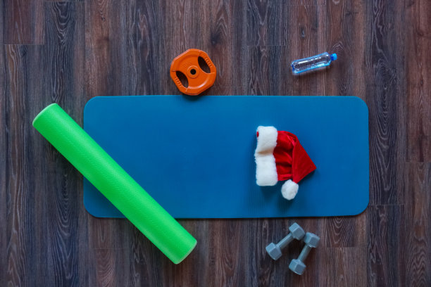 俯视图两个哑铃，圆盘，一瓶矿泉水，卷起的绿色垫子和圣诞老人的帽子躺在旁边的蓝色瑜伽垫子。健康的生活方式概念。本空间图片下载