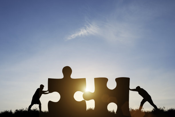剪影两个男人推动连接拼图与阳光和蓝天。它象征着企业团队合作的解决方案、成功和战略理念。图片下载