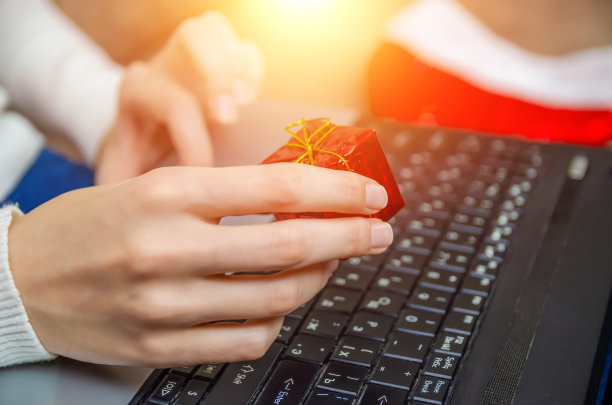 一个女人拿着一个红色的小礼盒在她的背景与一个圣诞老人的帽子，侧面的笔记本电脑。照片与照明。圣诞节、新年、节日的概念。图片下载