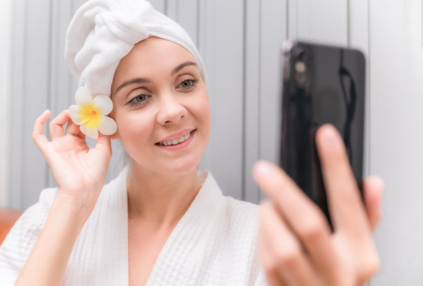 美丽的年轻女子在白色的连衣裙坐在沙发上，并通过手机自拍在水疗沙龙。图片下载