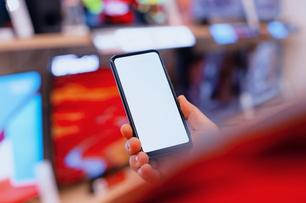 一个男人手里拿着一个白色屏幕的智能手机模型。商店里电视背景上的手机。图片下载